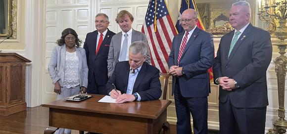 Picture of NC Governor Josh Stein, seated, signing House Bill 67 into law surrounded by people looking on