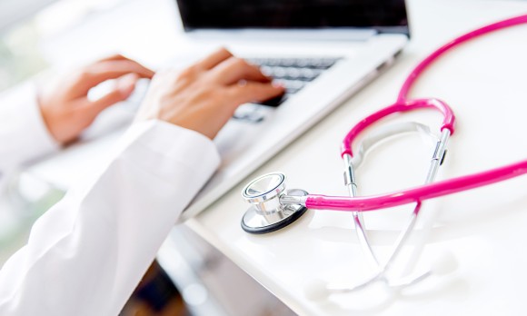 Doctor working on a laptop with a pink stethascope nearby on table